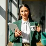 A woman in green blazer holding a mug with Baden Bower logo and a tablet smiling.