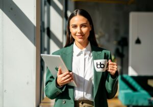 A woman in green blazer holding a mug with Baden Bower logo and a tablet smiling.