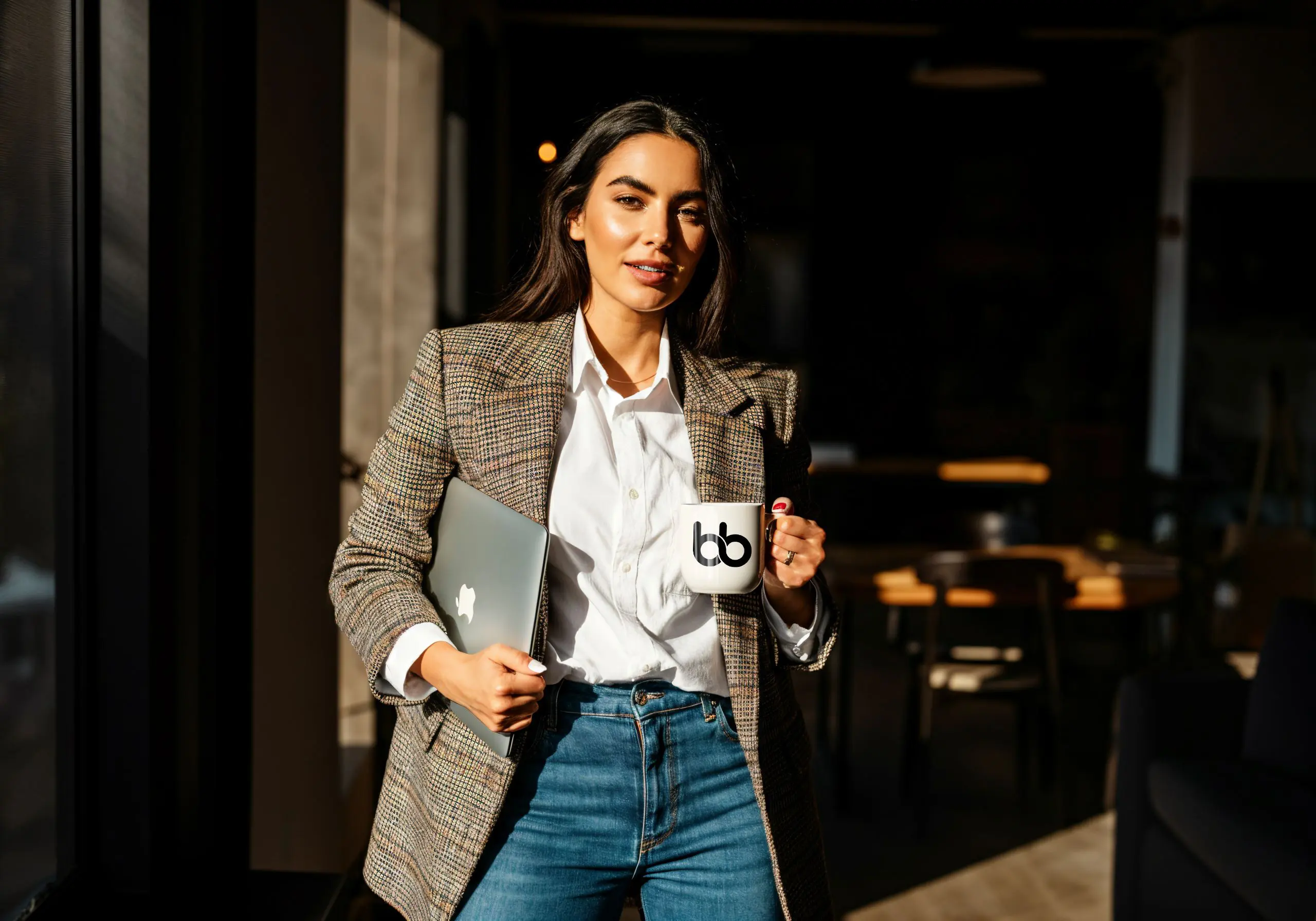 A woman in white shirt and a gray blazer holding a mug with Baden Bower logo on one hand and a laptop on the other.
