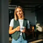 Professional in a green plaid vest and white tee holding a branded mug in a bright workspace, projecting a polished, magazine-ready look, get featured in Forbes.