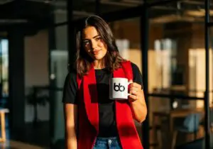 Professional in a bold red vest and black tee holding a branded mug near glass-walled meeting rooms, exuding confident, fashion-forward style, get featured in Vogue.