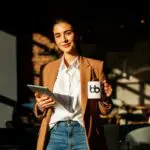 Professional in a camel blazer and white shirt holding a tablet and branded mug in dramatic sunlight, confidently ready for a presentation.